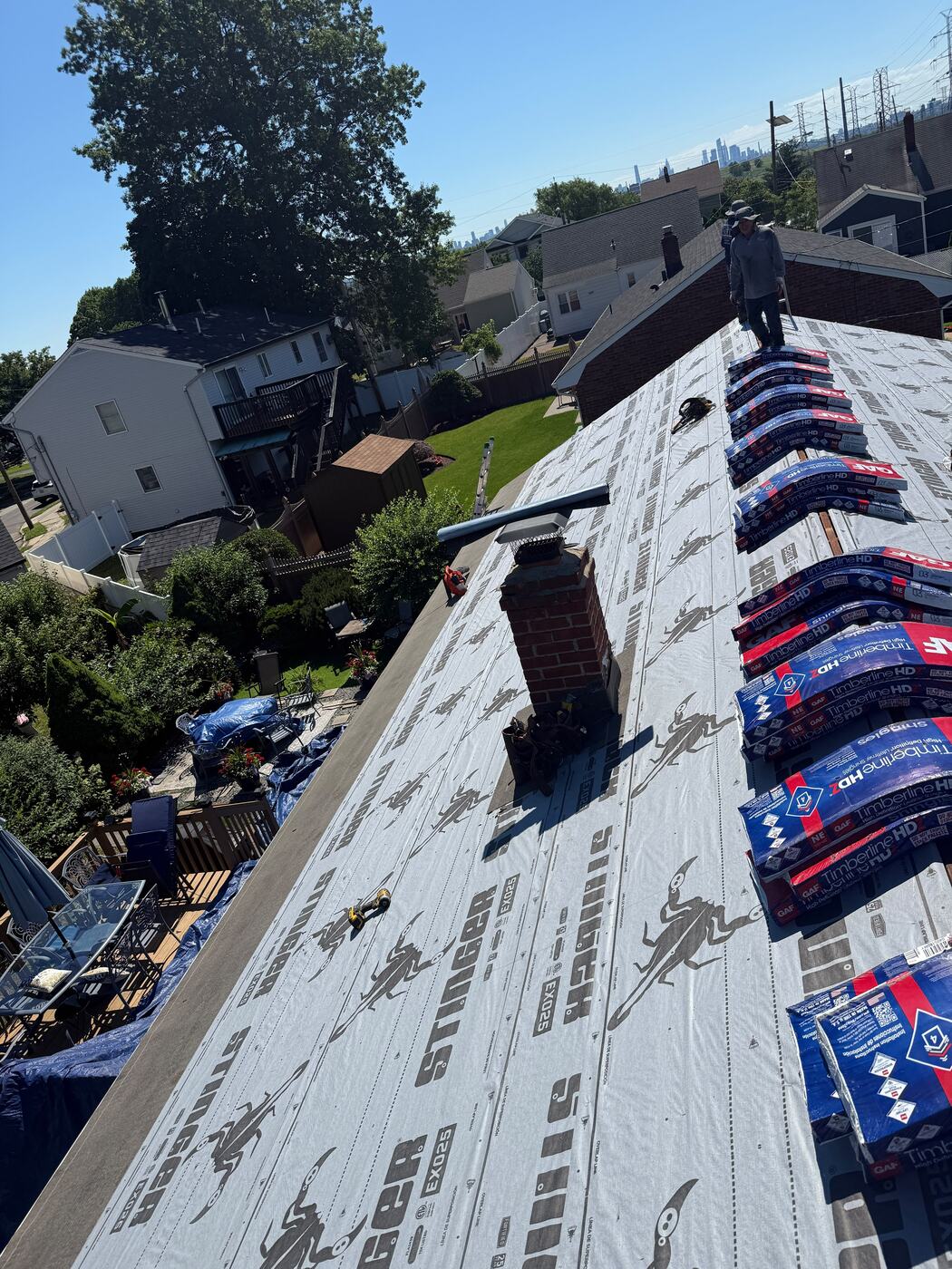 Brick chimney surrounded by new synthetic underlayment with shingle bundles staged along the ridge